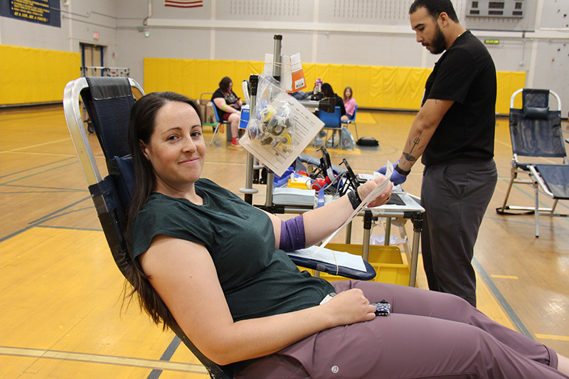 A woman with dark hair smiles. She is sitting with legs up on a cot after donating blood.