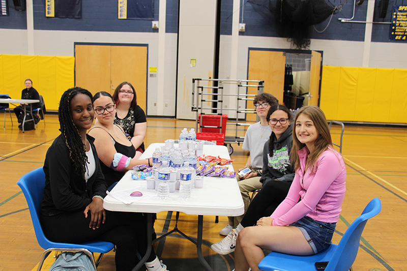 A group of six high school students sit on either side of a table that has snacks and drinks on it.