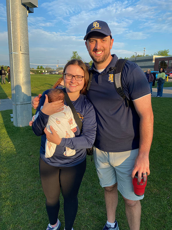 A tall man wearing a baseball style cap and a blue polo shirt that says PB on it smiles. He is standing wit a woman with hair pulled back and wearing glasses. She is smiling and holding an infant in her arms.