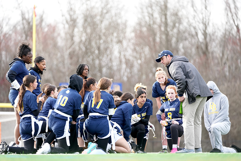 A man on the right bends in to talk to a team of high school girls flag football players. The are huddled up and wearing navy blue uniforms.