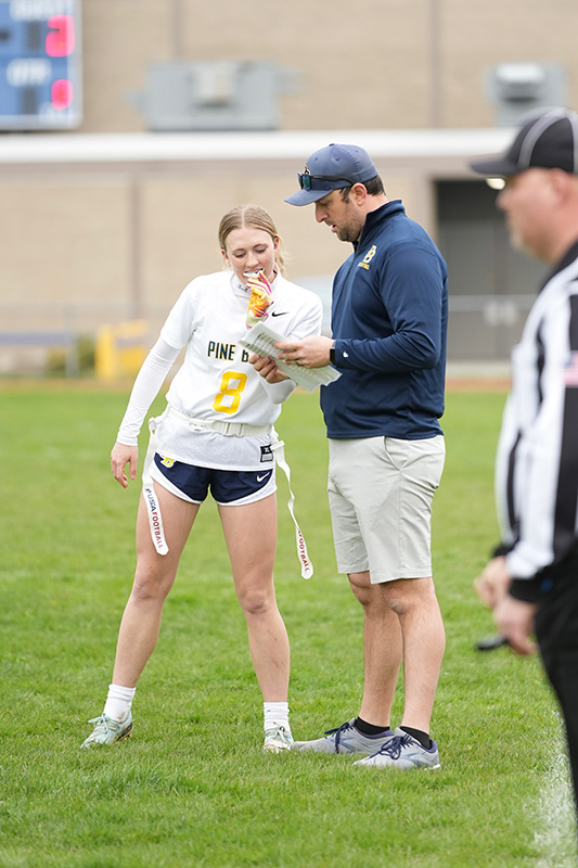 A tall man wearing a navy blue baseball type cap, navy shirt stands on the sidelines of a football field talking to a flag football player wearing a white Pine Bush shirt. Her hair is pulled back and she is looking at a piece of paper he is showing her.