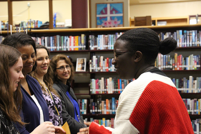 A high school junior dressed in a red and white shirt with hair in a ponytail shakes hands with a woman who hands her an envelope. Other adults are smiling and waiting to shake her hand.