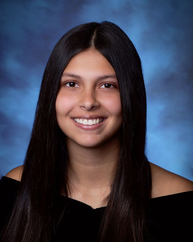 A young woman smiles. She has long dark hair and is wearing an off the shoulder black drape.