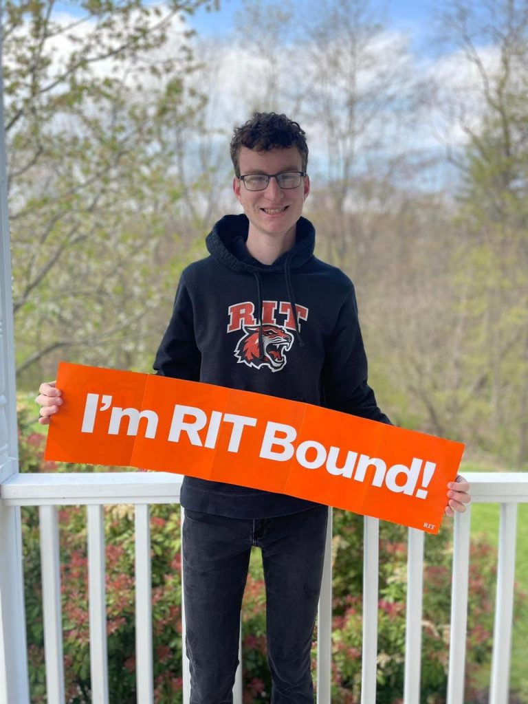 A high school senior boy with short hair and glasses. He is wearing a black hoodie  and holding a sign that says I'm RIT Bound. He is smiling.