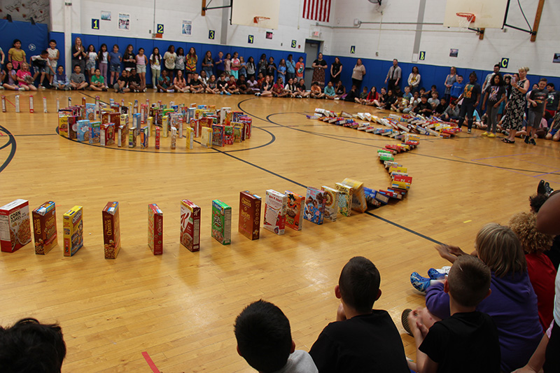 Hundreds of cereal boxes lined up and are falling into each other in a domino effect. Kids are sitting smiling and watching.