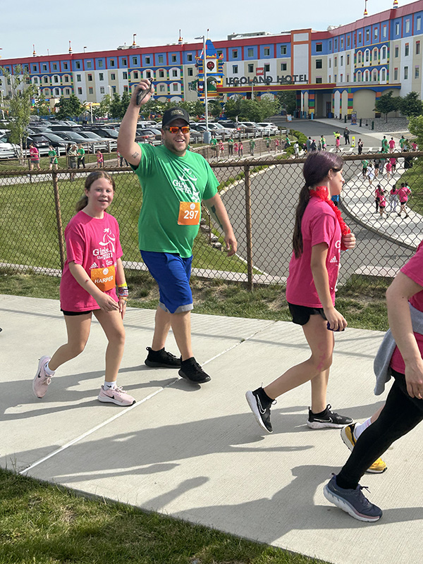 A man in a green shirt and baseball caps waves as a girl with a pink shirt runs with him.