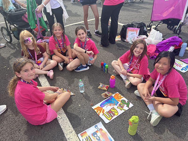 A group of six elementary girls all wearing pink tshirts sit on the ground together. and look up at the camera smiling.