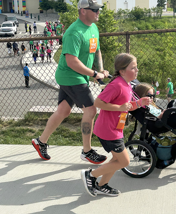 A man wearing a green tshirt pushes a jogging stroller. A girl wearing a pink tshirt runs along side of him and the stroller.