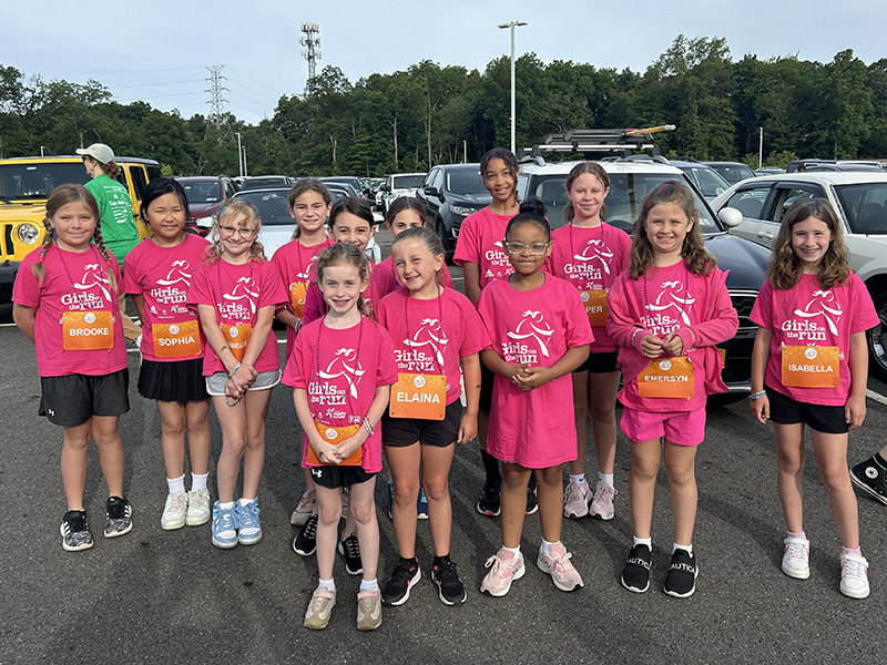 A group of about 12 elementary age girls, all wearing pink Girls on the Run tshirts, stand together and smile.