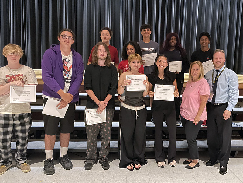 A group of 10 high school seniors standin gin two rows, along with a man and a woman, both on the right. The students are smiling and holding certificates.