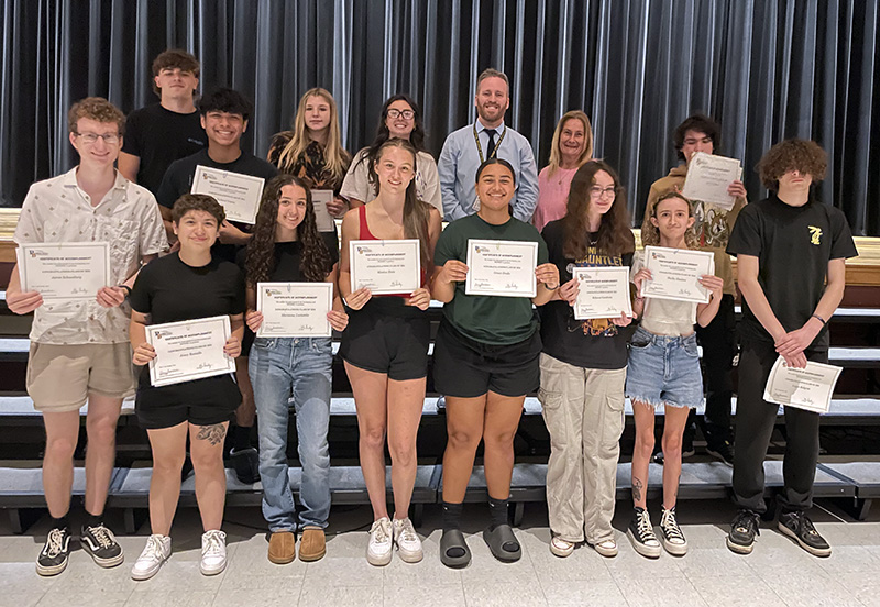 A group of 13 high school seniors smiling and holding certificates. They are standing in two rows with two adults, a man and woman. 