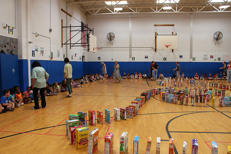 A gymnasium filled with cereal boxes in a wavy pattern. People are lined up and kids are sitting waiting for it to begin the domino.