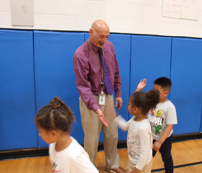 A man wearing a maroon shirt and blue tie smiles as he high fives smaller kids as they leave.