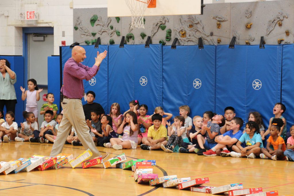 Younger elementary students sit along a blue wall waving and clapping. A man walks past them clapping.