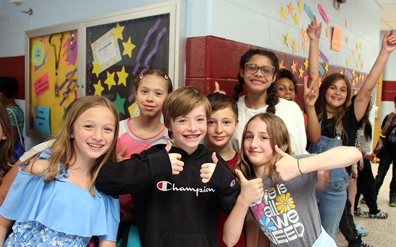 A group of eight fourth-graders smile and give thumbs up in the hallway of a school.
