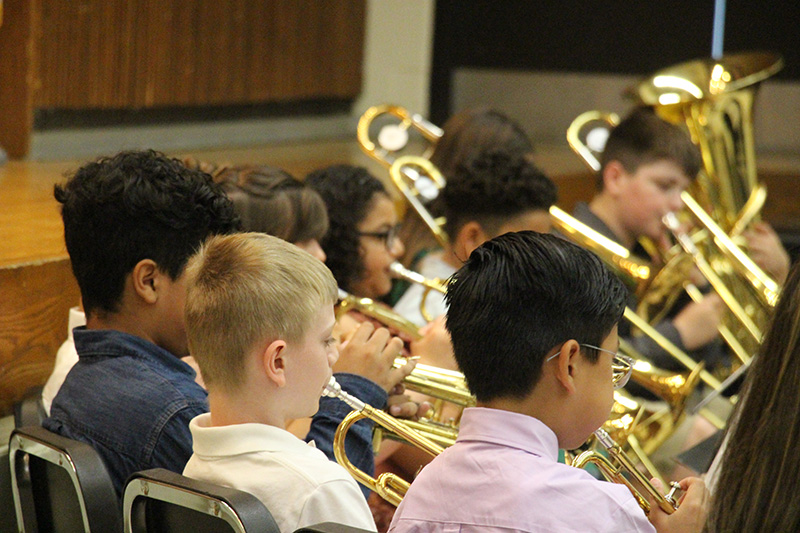 A group of fifth-grade students sit playing brass instruments.