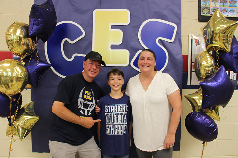 A background of blue with the letters CES in blue and gold. A man and woman flank a fifth-grade boy who is wearing a shirt that says. Straight outta fifth grade. The man is pointing to the shirt.