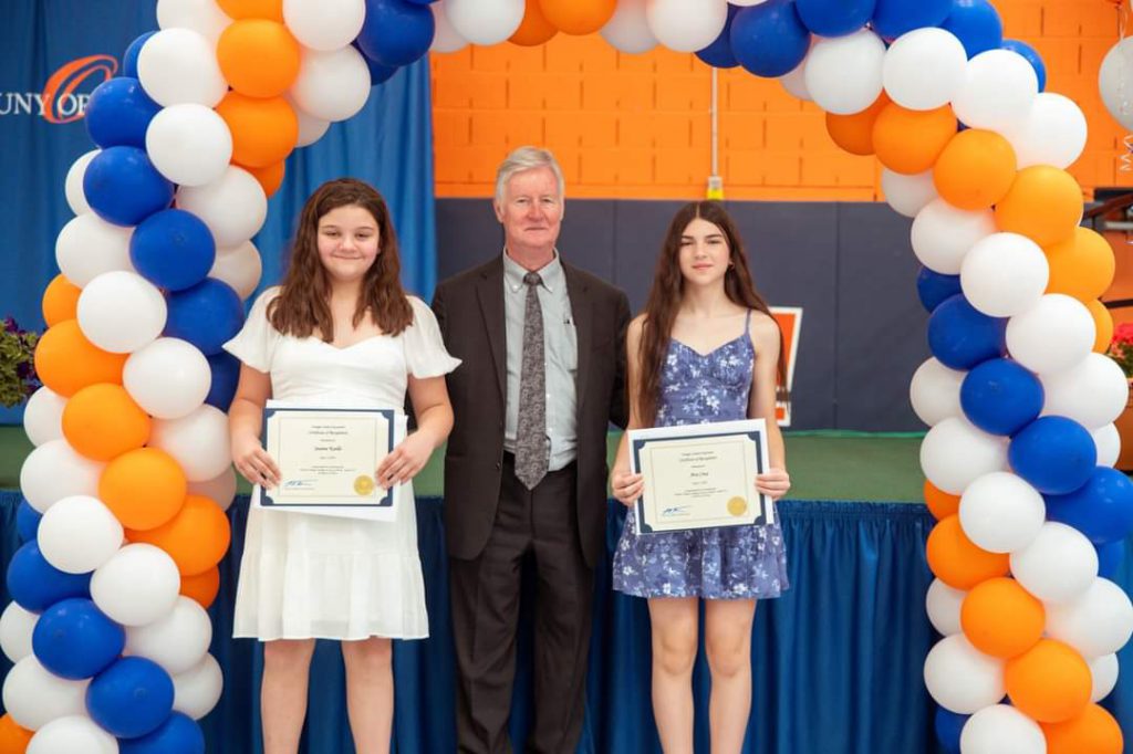 Two young women hold certificates and smile. There is a man in the center of them.