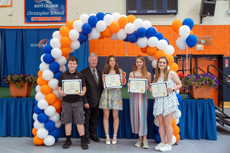 Three young women and a young man stand holding certificates under a blue, white and orange arch of balloons. With them is a man.