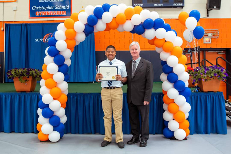 A young man holds his certificate and stands with a man. they are under a blue, white and orange balloon arch.