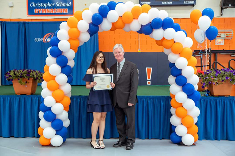 A young woman holds her certificate and stands with a man. They are under a blue, white and orange balloon arch.