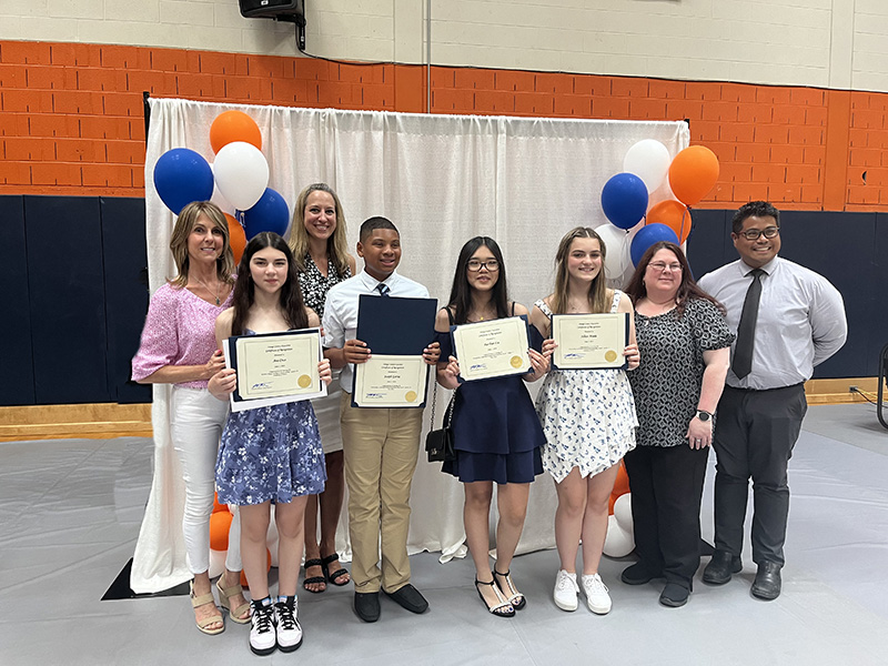 Four middle school students, one boy and three girls, hold up certificates and smile. There are four adults with them, two women on the left, and a woman and man on the right. There are blue, orange and white balloons.