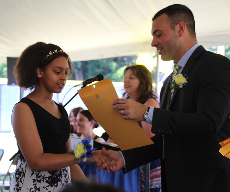 A fifth-grade girl wearing a black and white dress accepts a certificate from a man in a suit.