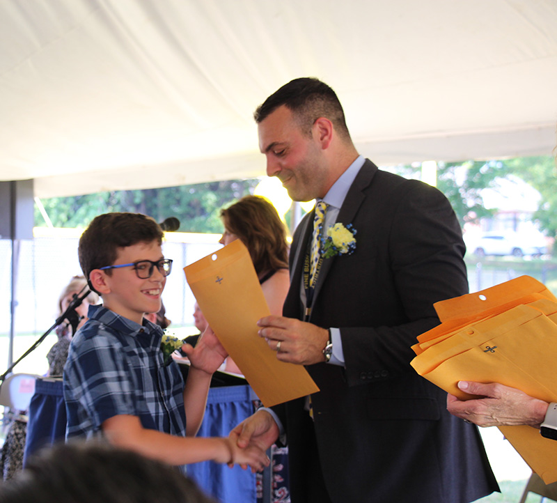 A man shakes a fifth-grade boys hand and hands him an envelope with a certificate. The boy has short dark hair and is wearing a plaid shirt and glasses.