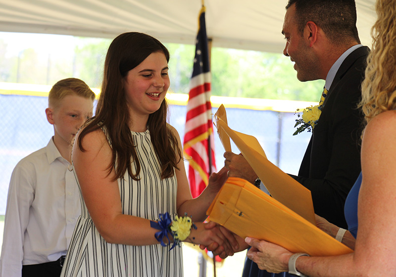 "A fifth-grade girl wearing a blue and white striped dress with long brown hair accepts a certificate from a man who is smiling.