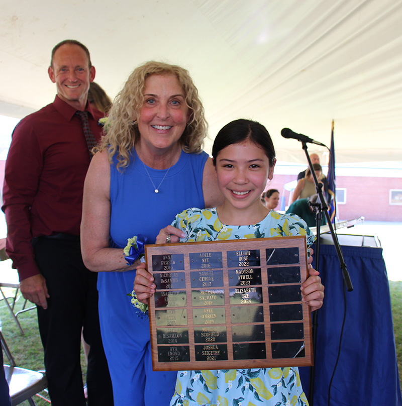 A fifth-grade girl with dark hair pulled back holds a large plaque with names engraved on it. Behind her is a woman with long blonde hair in a blue jumpsuit smiling as she leans in to the girl. Behind them is a man smiling.