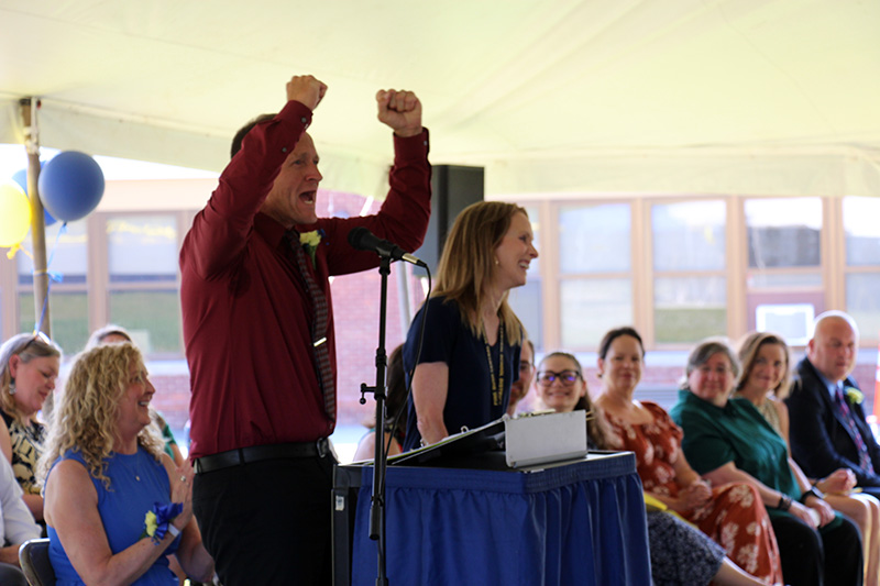 A man wearing a maroon shirt speaks at a podium. His hands are raised joyously. Next to him is a woman smiling.