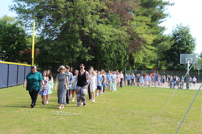 A long line of well-dressed fifth-grade students process across grass.