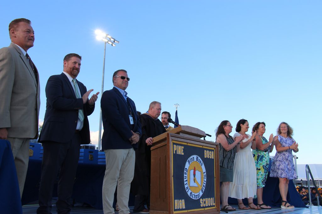 A blue sky in the background. A podium in the center of a stage with a man speaking. On either side are three men and four women. All clapping