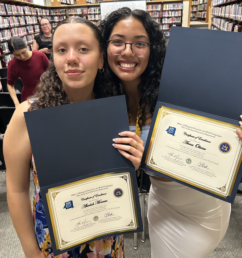 Two high school girls have their heads close together and smile. They are holding certificates.