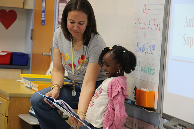 A woman with shoulder-length dark hair holds a book while a girl with a pink sweater and dark hair smiles and reads.