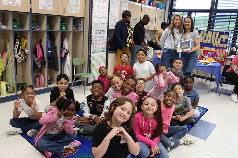 A group of about 20 second graders sit on a floor and smile for the picture. There are two women standing behind them, also smiling.