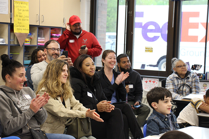 A group of parents sit in a classroom clapping.