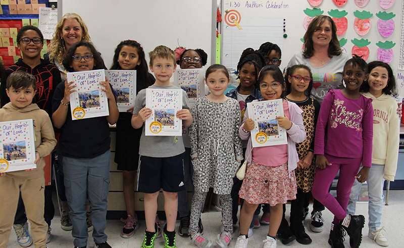 A group of about 20 second grade students stand in two rows, each holding up a white hardcover book.