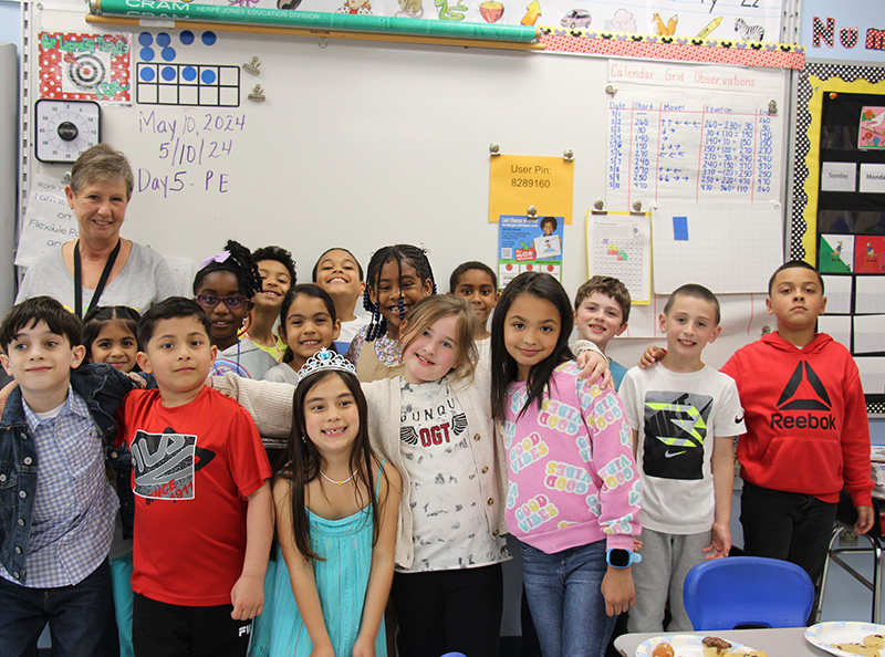 A group of about 20 second grade students and their teacher stand in front of a large white board. they are smiling.