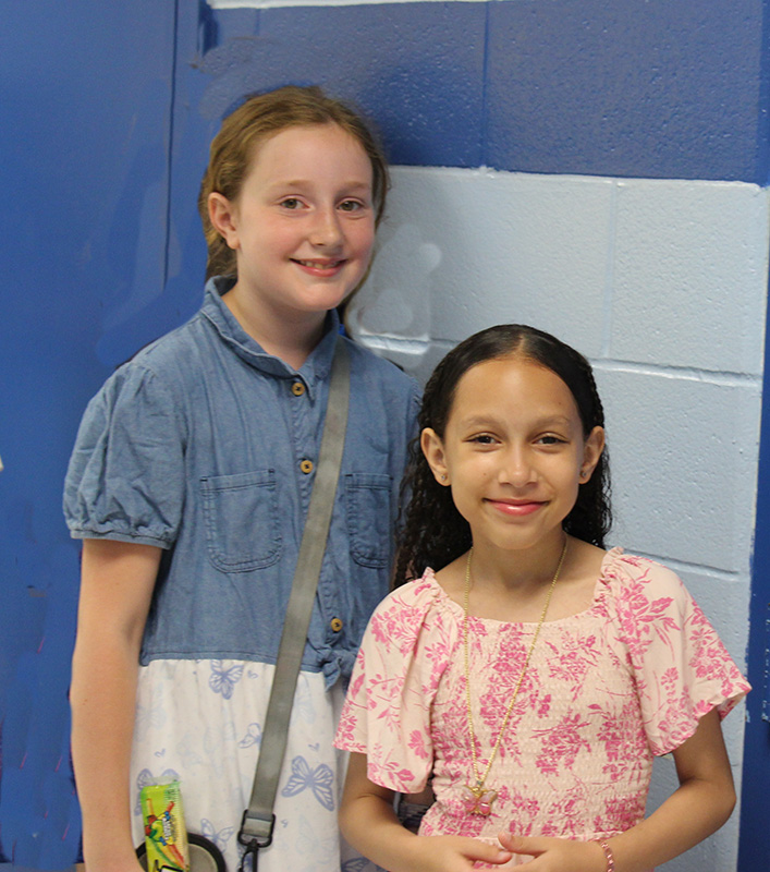 Two fifth-grade girls stand with each other smiling.