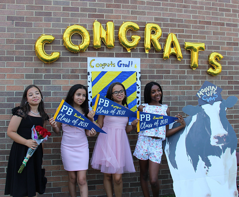 A group of four fifth-grade girls stand  by the congrats grad sign under gold balloons tha tspell out CONGRATS. There is a cutout of a cow next to them that says Moovin' up to CVMS.
