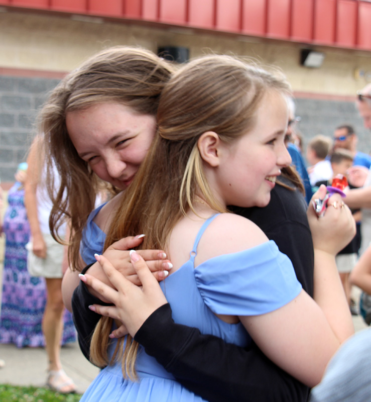 A fifth-grade girl with long blonde hair wearing a blue dress smiles as she hugs a woman.