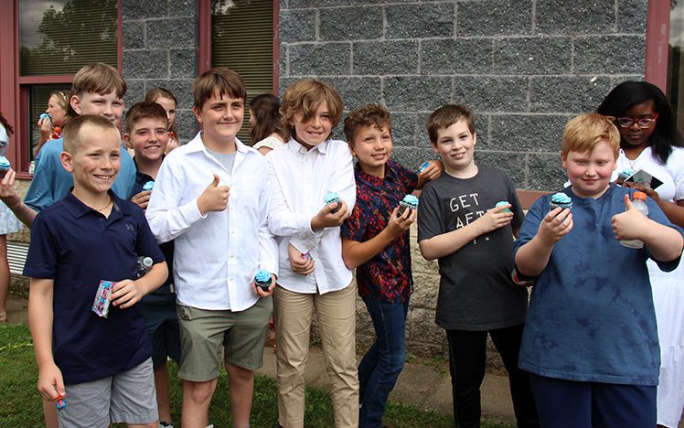 A group of seven fifth-grade boys stand together outside of a building. They are all smiling. Some are holding cupcakes.