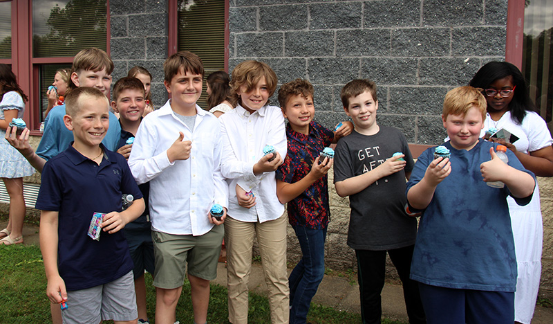 A group of seven fifth-grade boys stand together outside of a building. They are all smiling. Some are holding cupcakes.