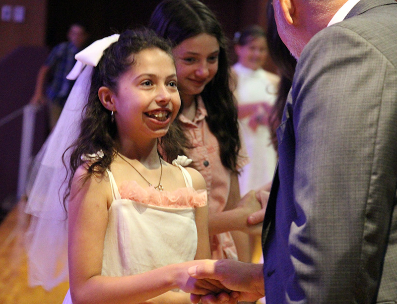 A fifth-grade girl wearing a white dress and a bow with a veil in her hair, smiles and shakes the hand of a man in a suit.