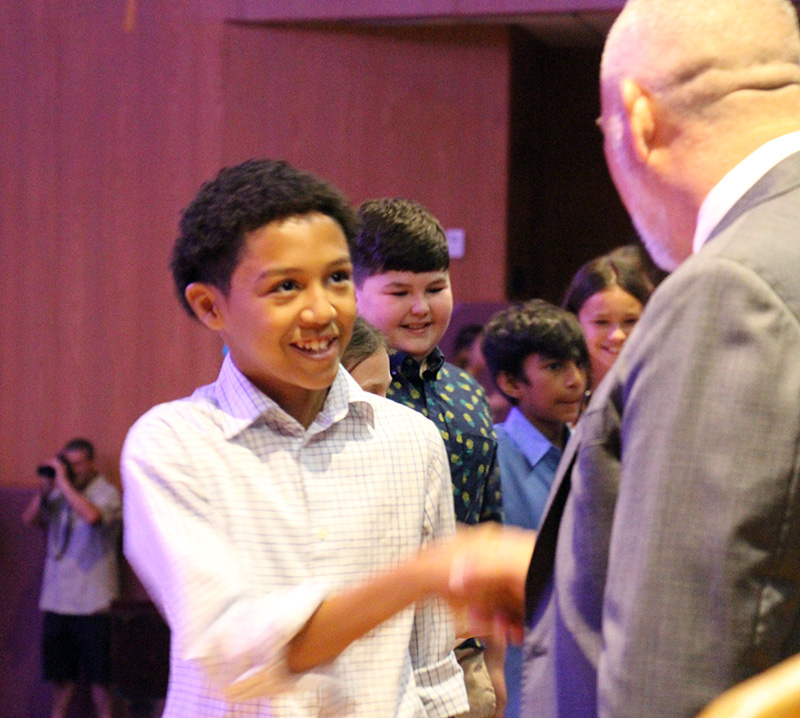 A boy with short dark hair smiles as he shakes the hand of a man in a suit.