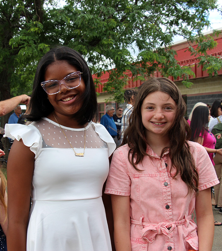 Two fifth=grade girls, dressed nicely, smile outside of a building.