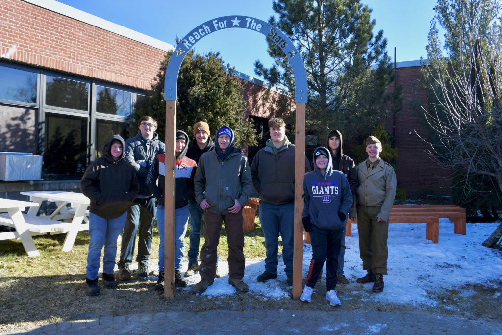 Nine high school students, dressed in winter coats and hats, stand together on a bright winter day. Snow on the ground. They are standing by an arch made of wood and metal they erected. The arch says Reach for the Stars.