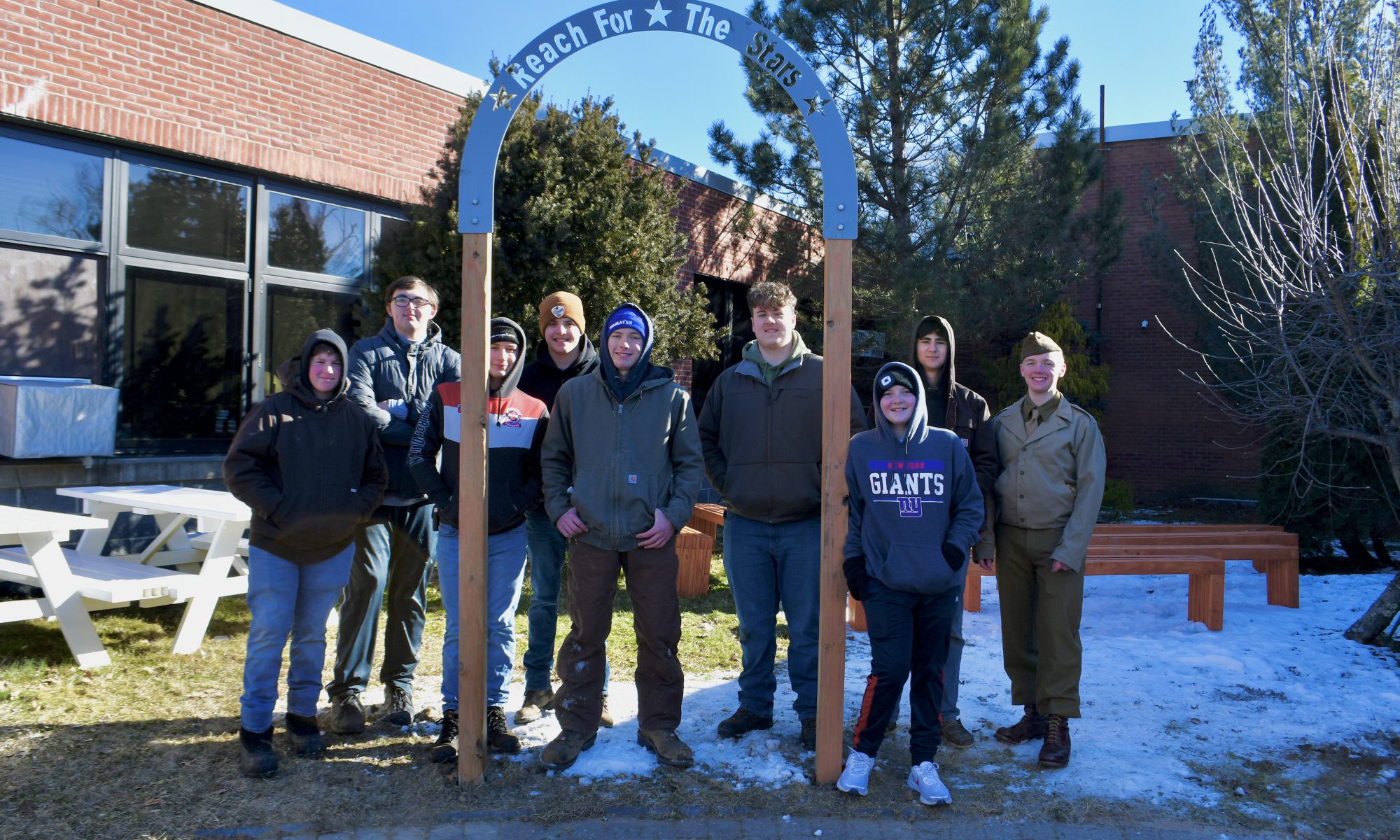 Nine high school students, dressed in winter coats and hats, stand together on a bright winter day. Snow on the ground. They are standing by an arch made of wood and metal they erected. The arch says Reach for the Stars.