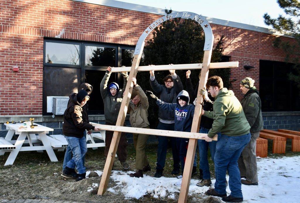 A group of nine high school boys hoist up an arch built from wood and metal. There is snow on the ground and they are dressed in winter coats.